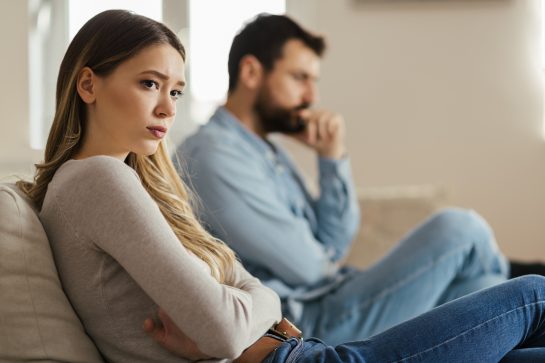 A woman sitting on a couch, she seems worried and looking away from her husband. Representing how one can benefit from calling a Naperville divorce attorney.