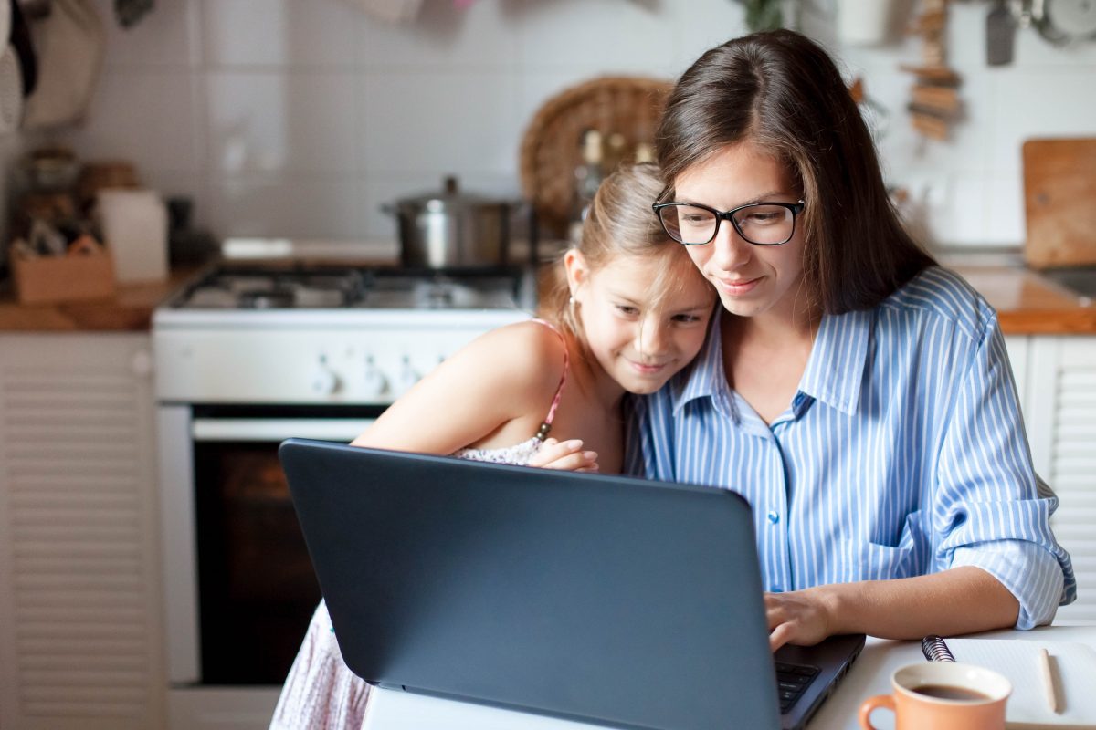 A woman wearing glasses sitting at a kitchen table wit her daughter looking at a laptop, representing how one can benefit from calling a Chicago child time-sharing attorney.